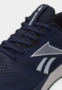 Αθλητικό παπούτσι Reebok σε ναυτικό μπλε με πλέγμα στην επιφάνεια, μπλε κορδόνια και λευκή λεπτομέρεια λογότυπου στο πλάι και στη γλώσσα, εμφανιζόμενο σε κοντινή γωνία.