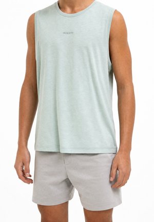 Man met een lichtgroene mouwloze top met de tekst "Morato" en lichtgrijze korte broek, staand met ontspannen armen langs het lichaam.