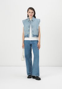  colete de denim com design sem mangas, na cor azul clara, e botões na frente. Combinado com calças jeans largas azuis e uma pequena bolsa branca.