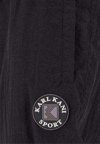 Juodos kelnės su tekstūruota apdaila, turinčios apvalų gumos ženkliuką su užrašu "KARL KANI SPORT" balta ir pilka spalvomis.