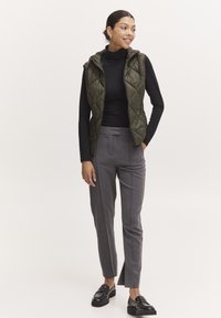 Gilet matelassé vert olive avec capuche, associé à un col roulé noir et un pantalon gris ajusté, avec des fentes latérales et des mocassins noirs.