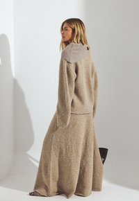 Completo lavorato a maglia di un colore marrone chiaro con cappuccio, caratterizzato da una superficie testurizzata. Maniche lunghe e ampie e una gonna che arriva fino al pavimento. Accento del cappuccio in grigio liscio.