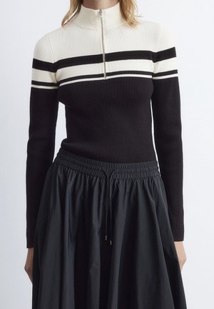 Femme portant un pull côtelé noir et blanc à rayures horizontales avec une fermeture éclair à l’avant, associée à une jupe noire froncée avec un lien de serrage.