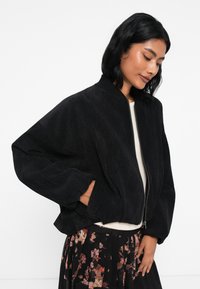 Svart kordfløyelsjakke med kort design, ribbede mansjetter og glidelås foran. Kombinert med et blomsterkjørt og en kremfarget topp.