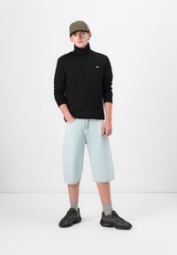 Un pull à col roulé noir, un short en denim bleu clair jusqu'aux genoux, des chaussettes grises, des chaussures de sport noires et une casquette à carreaux bruns.