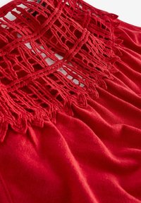 Robe rouge avec un corsage en dentelle crochetée texturée aux motifs délicats et un tissu doux et drapé. Le tissu semble lisse et léger.