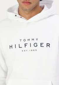 Biała bluza z kapturem wykonana z bawełny, z kieszenią kangurką i ściągaczami. Na przodzie nadrukowany czarny napis "TOMMY HILFIGER EST.1985".