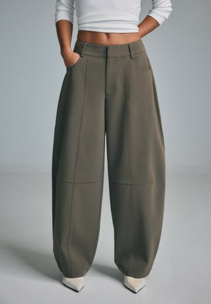 Person wearing high-waisted wide-leg olive green trousers with hands in pockets and white pointed shoes, cropped white long sleeve top.