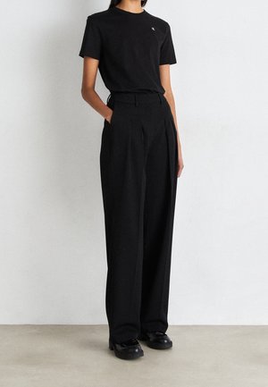 T-shirt noir en coton à manches courtes avec logo sur la poitrine, assorti à un pantalon noir ajusté avec une coupe ample et des plis. Chaussures noires.