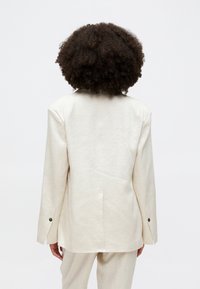 Beige Leinenblazer mit einem einzelnen Rückenventil, taillierte Passform, langen Ärmeln und schwarzen Knöpfen an den Manschetten, von hinten vor einem einfarbigen Hintergrund betrachtet.