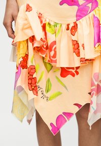 Jupe à imprimé floral en tissu pêche doux, avec un ourlet à volants et une coupe asymétrique. Fleurs rouge vif et rose avec des accents verts.