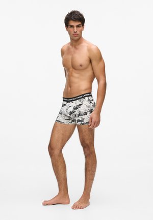 Krekla nesaturošs vīrietis, stāv basām kājām, valkājot baltas bokserbikses ar melnu tropisku rakstu un melnu jostasvietu ar uzrakstu Karl Lagerfeld.