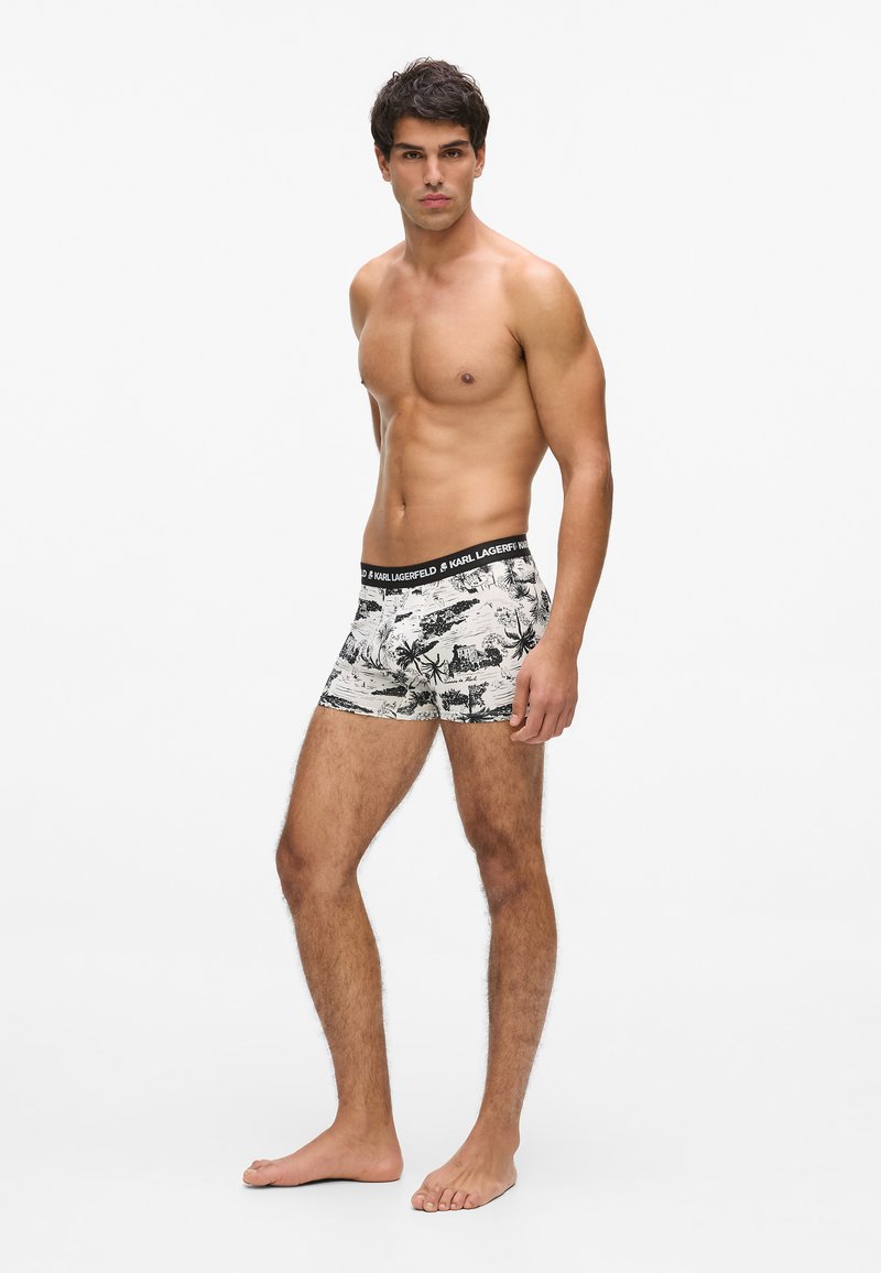 Muž bez trička stojaci na boso, oblečený v bielych boxerkách s čiernym tropickým vzorom a čiernym pásom s nápisom Karl Lagerfeld.