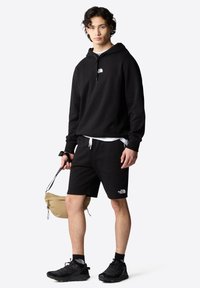 Sudadera negra con cordones, combinada con pantalones cortos negros. Presenta el logo de The North Face. El modelo sostiene una bolsa color marrón, vistiendo zapatos y calcetines negros.