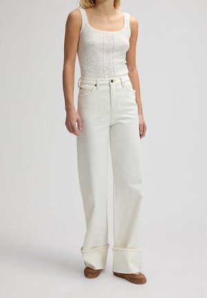 Mujer con top sin mangas texturizado blanco, pantalones blancos de pierna ancha y cintura alta con puños remangados, y zapatos planos marrones, de pie frente a un fondo liso.