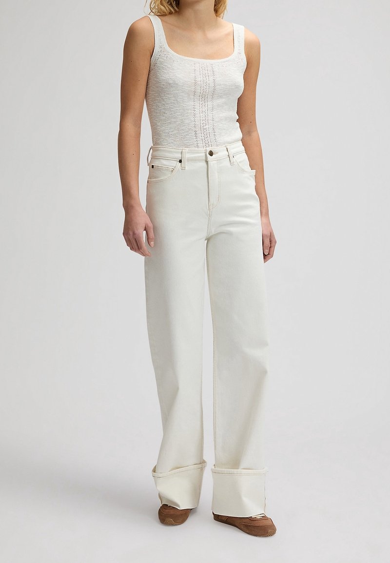 Femme portant un top sans manches blanc texturé, un pantalon blanc taille haute à jambes larges avec revers roulés, et des chaussures plates marron, debout devant un fond uni.