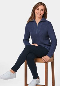 Marineblauer Reißverschluss-Pullover mit geripptem Design, kombiniert mit dunklen Jeans und weißen Sneakers. Das Model sitzt auf einem Holzschemel und lächelt.