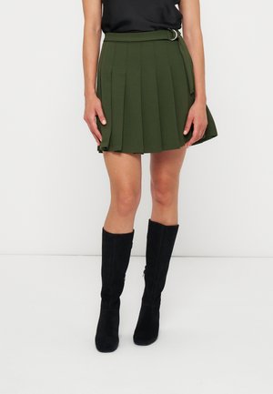 Femme portant une mini-jupe plissée vert olive avec une ceinture et des bottes noires montantes jusqu'au genou, debout sur un fond blanc.
