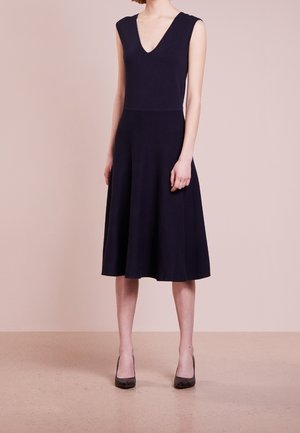 Femme portant une robe noire sans manches avec un col en V et une jupe évasée, debout sur un sol et un fond beiges unis.