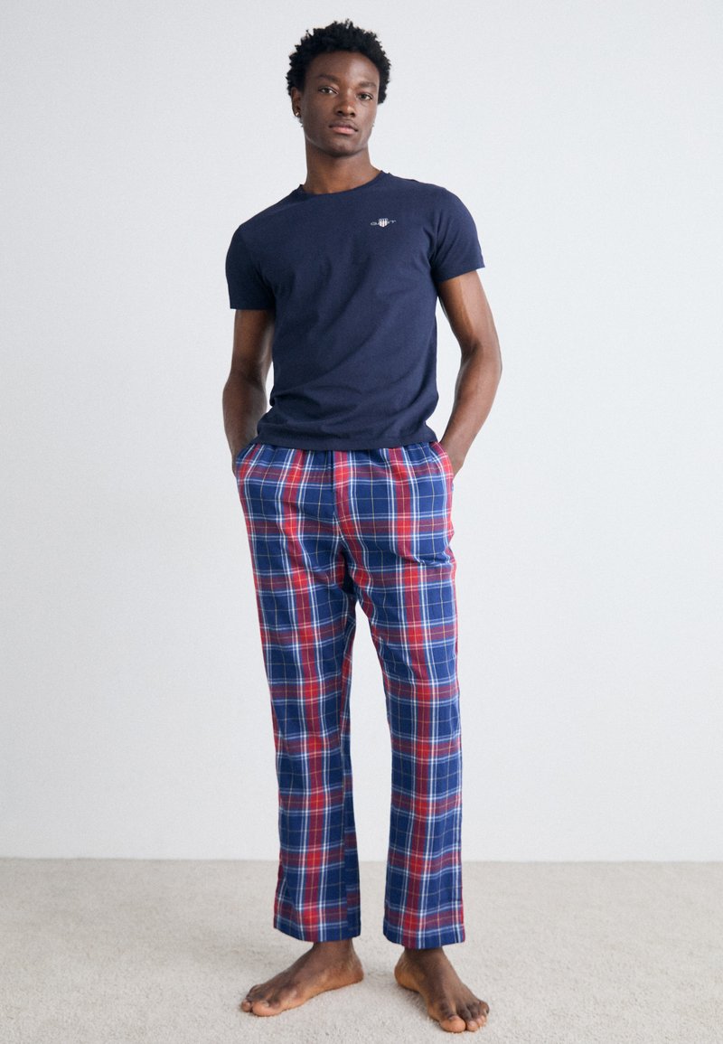 Marineblaues Baumwoll-T-Shirt mit kleinem Logo, kombiniert mit locker sitzenden Schlafanzughosen im rot-blauen Karomuster. Keine Schuhe, steht auf einem hellen Teppich.