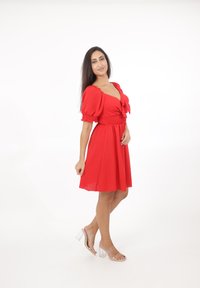 Rode jurk met pofmouwen en een strikdetail aan de voorkant. Vloeiende rok en elastische taille. Draagt met duidelijke sandalen met blokhak.