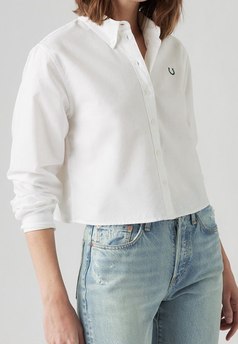 Chemise blanche courte avec devant à boutons, manches longues et logo brodé vert sur le côté gauche de la poitrine. Associée à un jean bleu clair déchiré.