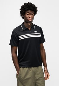 Μαύρο πόλο με μισό φερμουάρ και λευκές οριζόντιες ρίγες, με γιακά με σχέδιο και διακριτικό λογότυπο. Φοριέται με πράσινο σορτς.