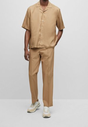 Homme portant une chemise à manches courtes beige boutonnée, un pantalon beige assorti et des baskets blanches, debout devant un fond gris uni.