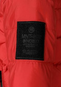 Giacca imbottita rossa con un'etichetta nera testurizzata con testo in rilievo che recita "MNT SINCE 97," cucita lungo la cucitura della manica.