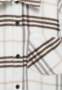 Camicia in flanella a quadri bianca e marrone con bottoni neri e tasca sul petto. Texture morbida e motivo a quadri con varie sfumature di marrone.