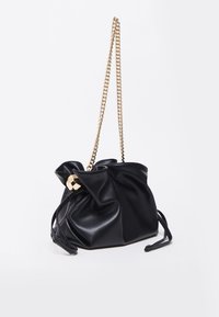 Sac en cuir noir à cordon avec bandoulière chaîne dorée et détail de fermeture hexagonale dorée sur fond blanc.