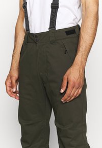 Olivgrön snöbyxor med justerbara svarta hängslen, med flera fickor, tryckknappsstängningar och förstärkt sömnad vid knäna.