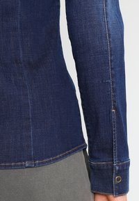 Denimhemd, dunkelblau, taillierte Passform mit einer Naht am Ärmel. Verfügt über einen Knopfverschluss am Bündchen mit einer Metallöse und kontrastierenden Nähten.