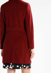 Bordový vlněný kabát s vysokým límcem, dlouhými rukávy a páskem v pase, s zadními švy a hladkým strukturálním povrchem.