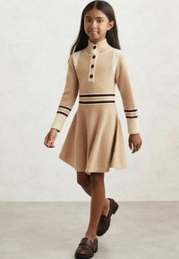 Dzianinowa sukienka w odcieniu jasnobrązowym z wysokim kołnierzem, zapięciem na guziki oraz paskami; rozszerzana spódnica; zestawiona z ciemno-brązowymi butami wsuwanymi.