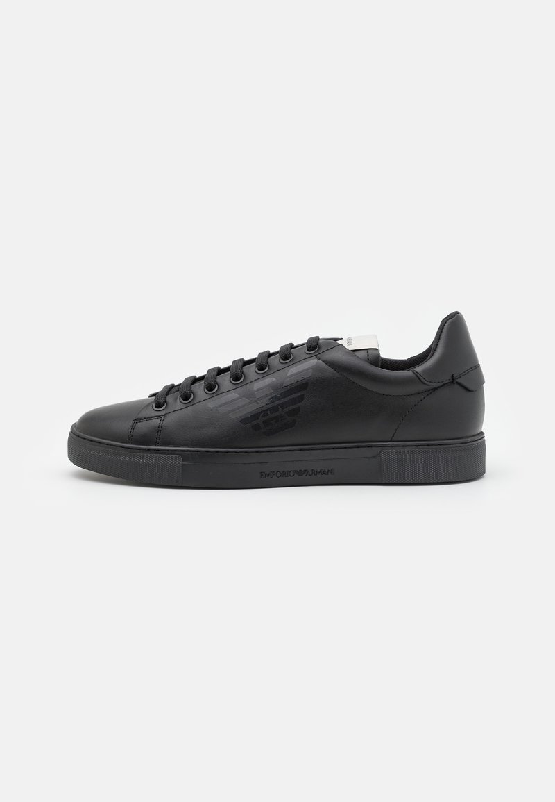 Baskets basses en cuir noir avec lacets noirs, logo embossé sur le côté et semelle en caoutchouc texturée sur fond blanc.