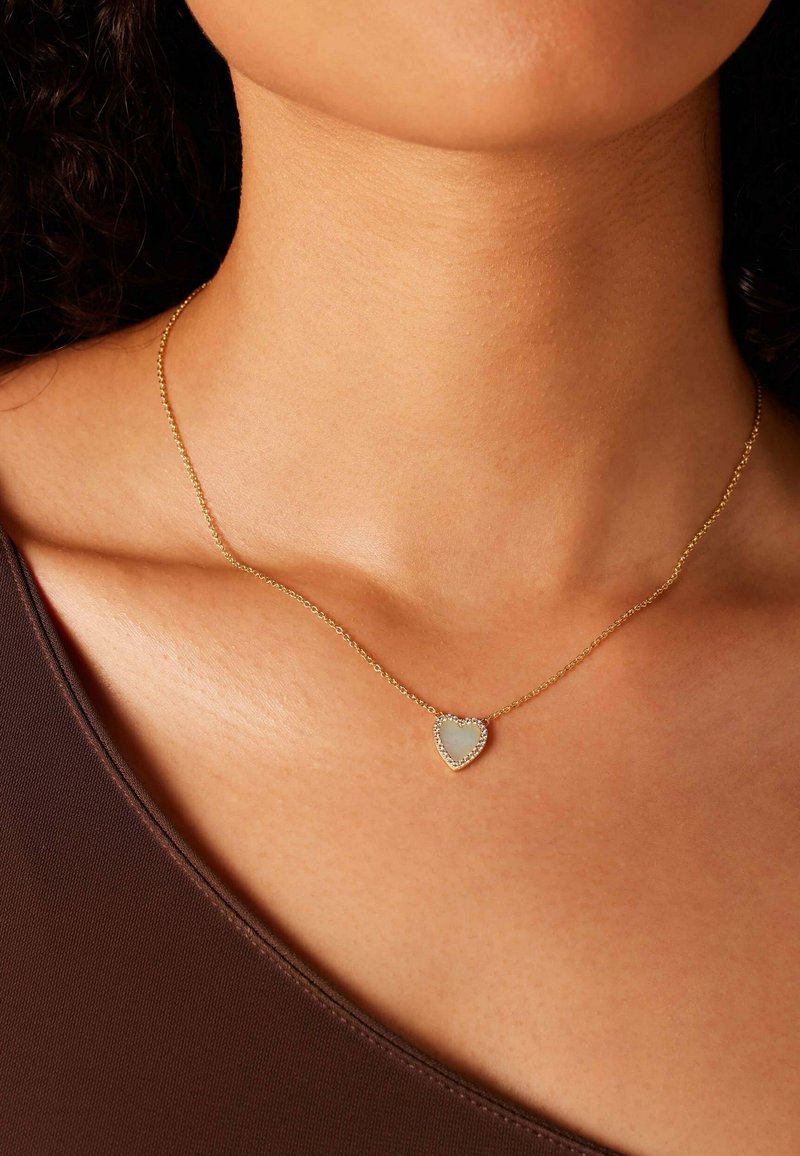 Gouden ketting met een hartvormige hanger met een licht ondoorzichtige center en een omliggende rand van kleine heldere stenen.