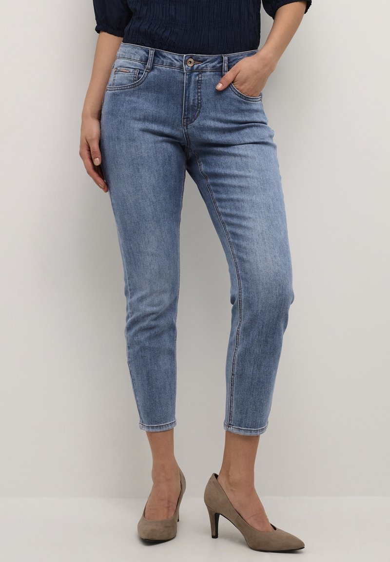 Femme portant un jean slim court bleu clair avec poches avant, associé à des escarpins beiges et un haut foncé.