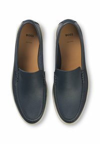 Marina läder slip-on skor med en slät yta, kontrasterande ljus sula och dekorativ sömnad längs de övre kanterna. Logotyp tryckt inuti.