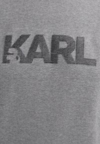 Tessuto grigio con lettere nere in rilievo che formano la parola "KARL" in un font audace e moderno, centrato sul materiale.