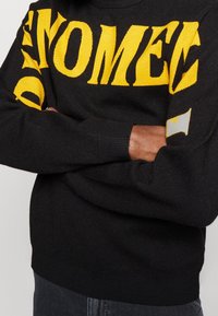 Pull noir avec lettrage jaune audacieux. Poignets et taille côtelés, texture douce, et coupe décontractée. Accent notable sur la manche gauche.