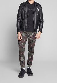 Chaqueta de motocicleta de cuero negra, camiseta negra ajustada, pantalones de camuflaje con puños elásticos y zapatillas negras con acentos blancos.