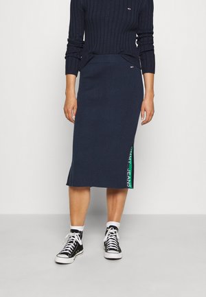 Femme portant une jupe côtelée marine avec le logo "Tommy Jeans" près de l'ourlet, un pull côtelé assorti marine, des chaussettes blanches et des baskets montantes noires.