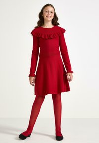 Punainen neulemekko, jossa on pitkät hihat ja röyhelökaulus, vartalonmyötäinen yläosa, levenevä hameosa, yhdistettynä punaisiin leggins-tai mustiin balleriinoihin.