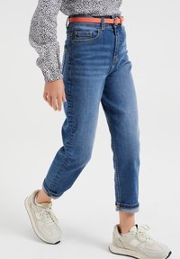 Džínové kalhoty s vysokým pasem, modré, rovného střihu, s ohrnutými manžetami a bočními kapsami, v kombinaci s světlými teniskami a růžovým opaskem.