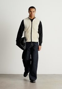 Μπεζ και μαύρο φλις γιλέκο με φερμουάρ πάνω από μαύρο μακρυμάνικο μπλούζα, συνδυασμένο με σκούρες φαρδιές τζιν παντελόνες και μαύρα παπούτσια, κρατώντας μια μαύρη τσάντα.