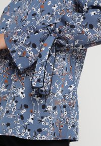 Tissu bleu avec un motif floral en blanc, noir et marron, avec un nœud noué sur une manche longue.