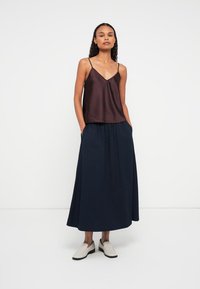 Kobieta w ciemnoszarej satynowej koszulce na ramiączkach, długiej granatowej plisowanej spódnicy z kieszeniami i białych butach wsuwanych, stojąca na tle jednolitego białego tła.