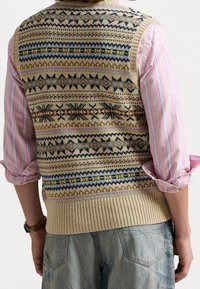 Beige stickad väst med färgglada geometriska mönster i blått, rosa och grönt, med en ribbad nederdel. Kombinerad med en rosa och vit randig skjorta.