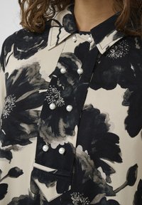 Blusa floreale nera e crema con colletto, dettagli in perle lungo il davanti e un tessuto texturizzato con una finitura liscia.